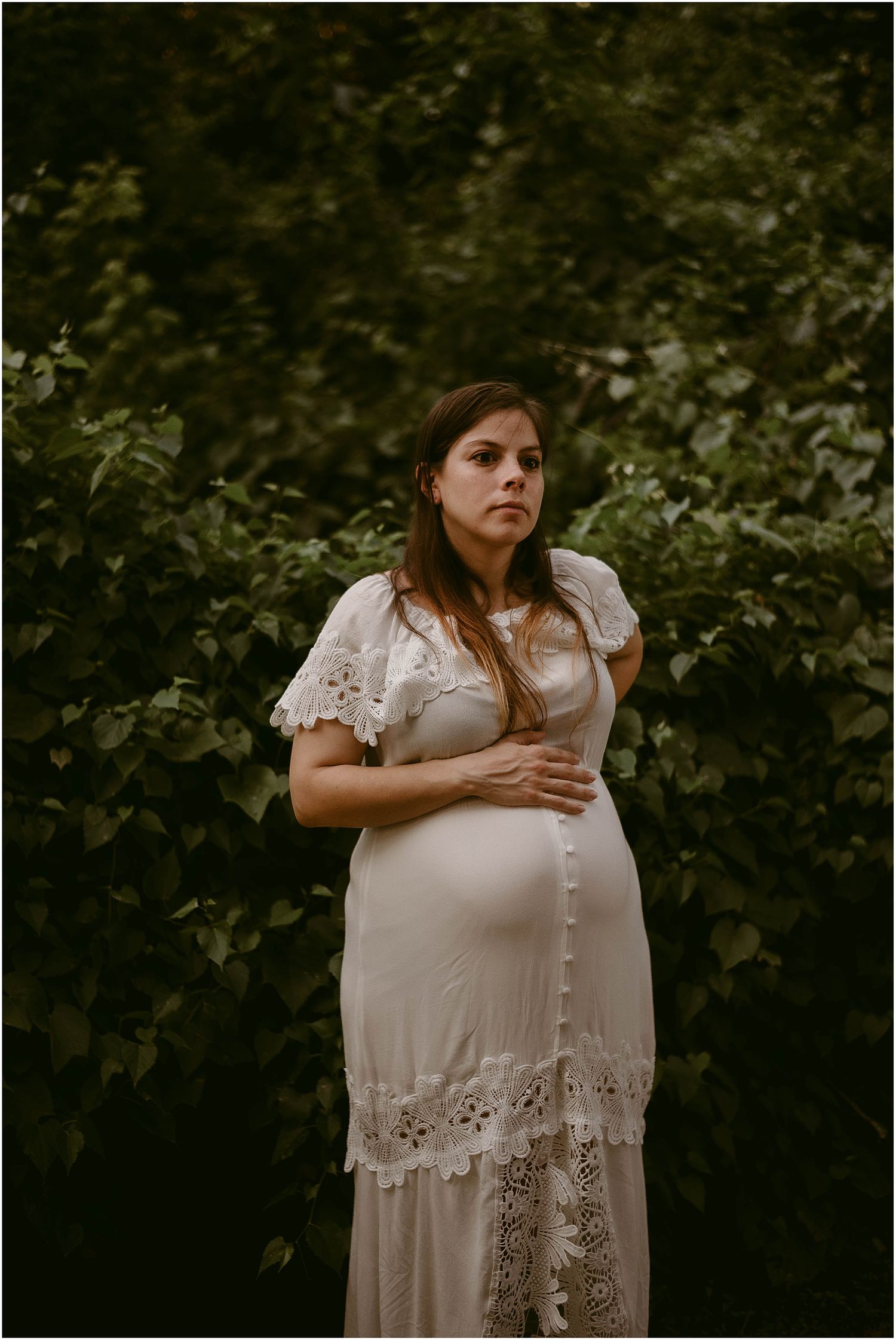 Elegant maternity portrait featuring intricate lace dress against dark green foliage.