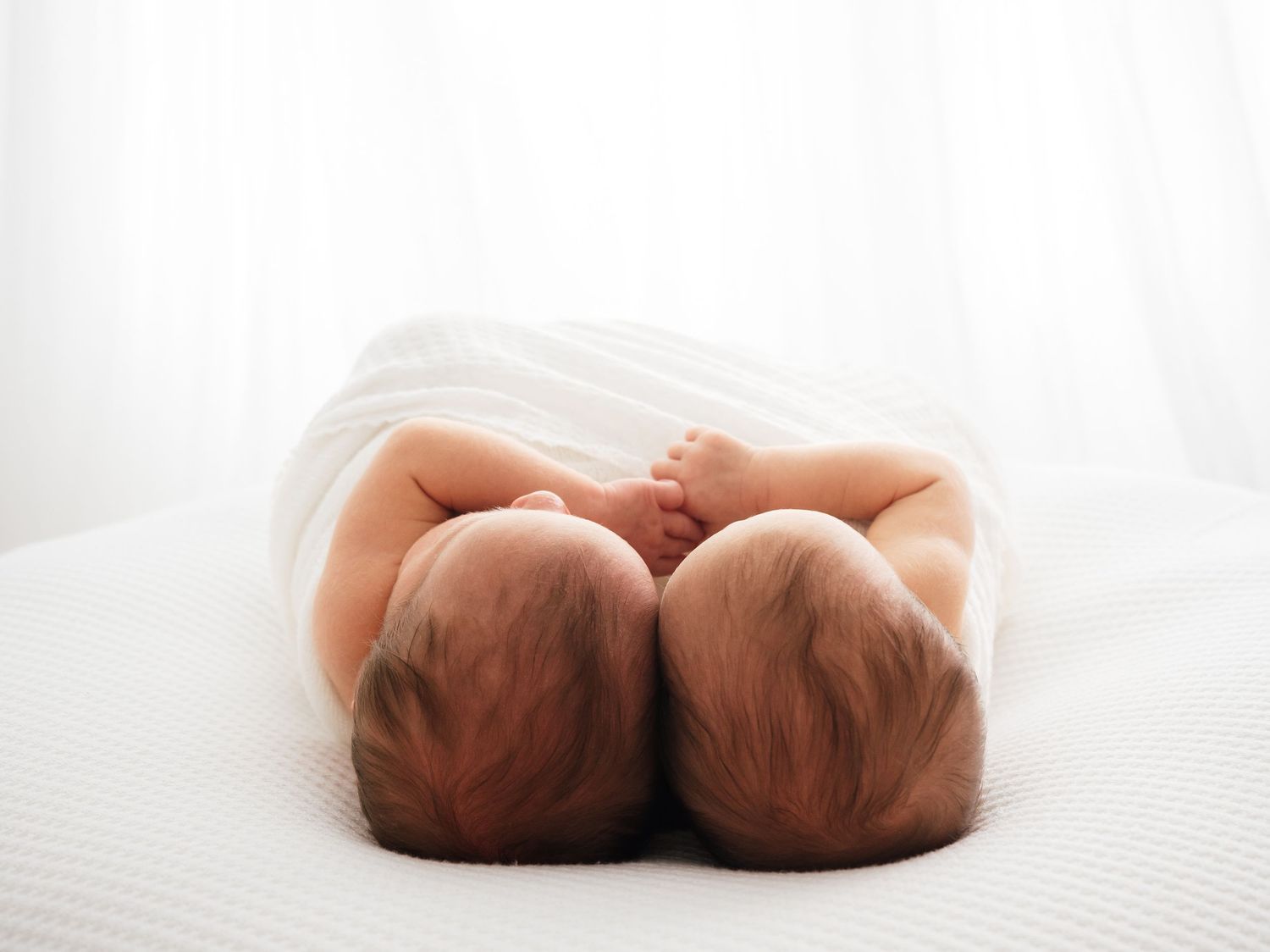 Photo of newborn twins swaddled in a white blanket and holding hands. By Verity Photography in Didcot