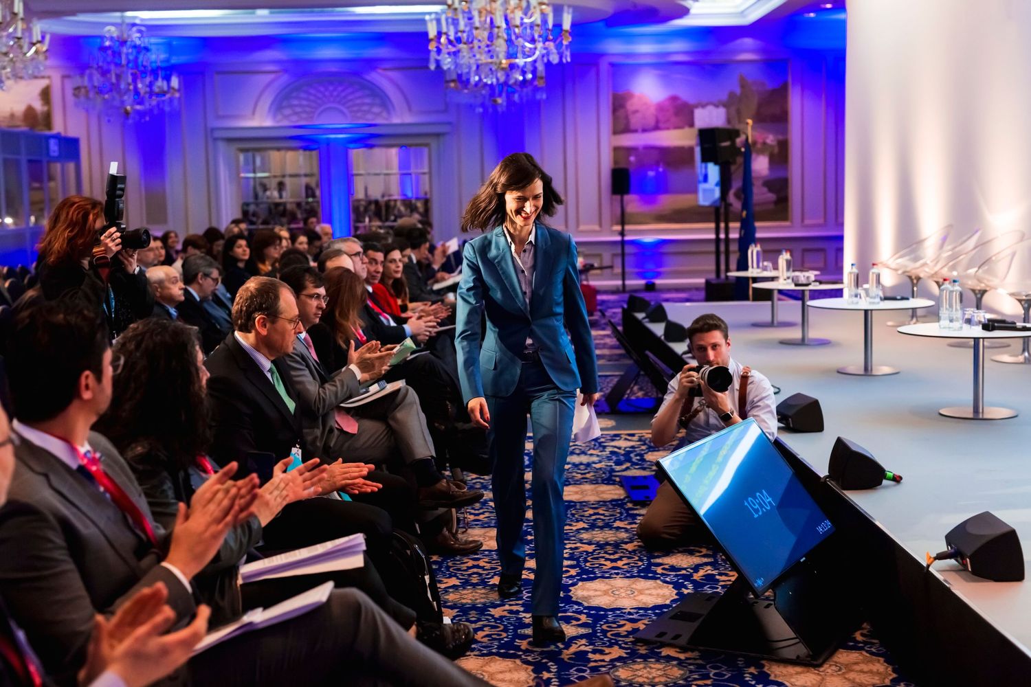Speaker presenting to an audience in an elegant ballroom with blue lighting and chandeliers.