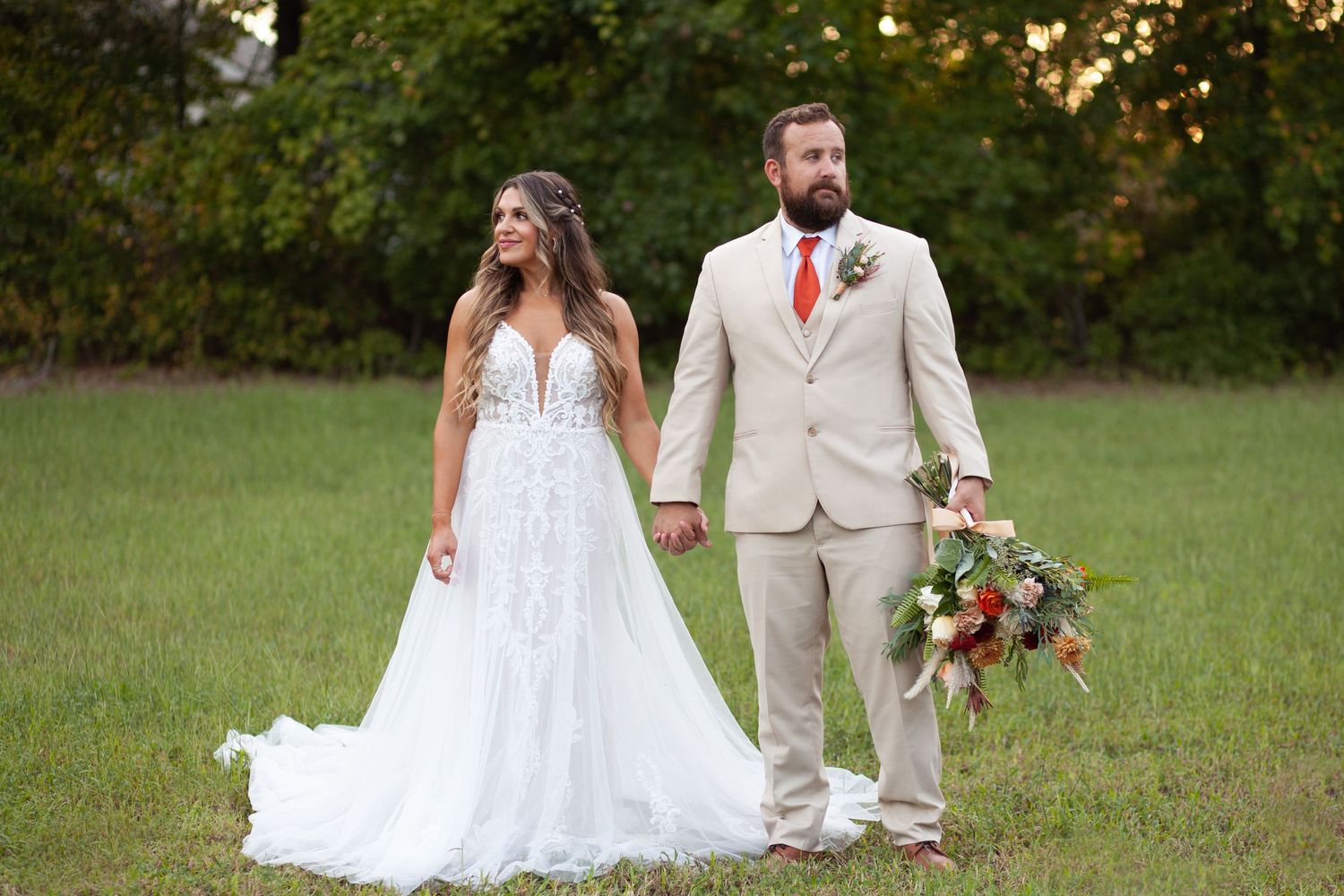A couple in wedding attire stands together in a grassy field at sunset while holding a colorful autumn bouquet at the House on Barber Mill.