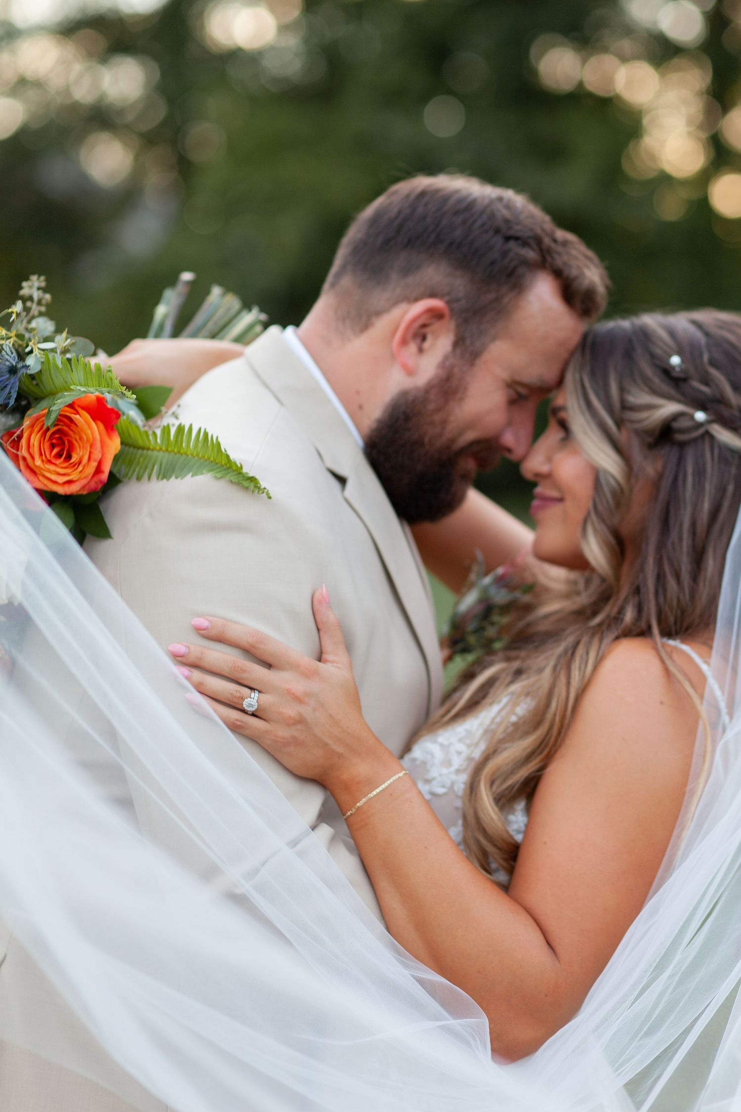 A romantic wedding couple embraces outdoors, with the bride in white and groom in light beige suit with orange boquet.