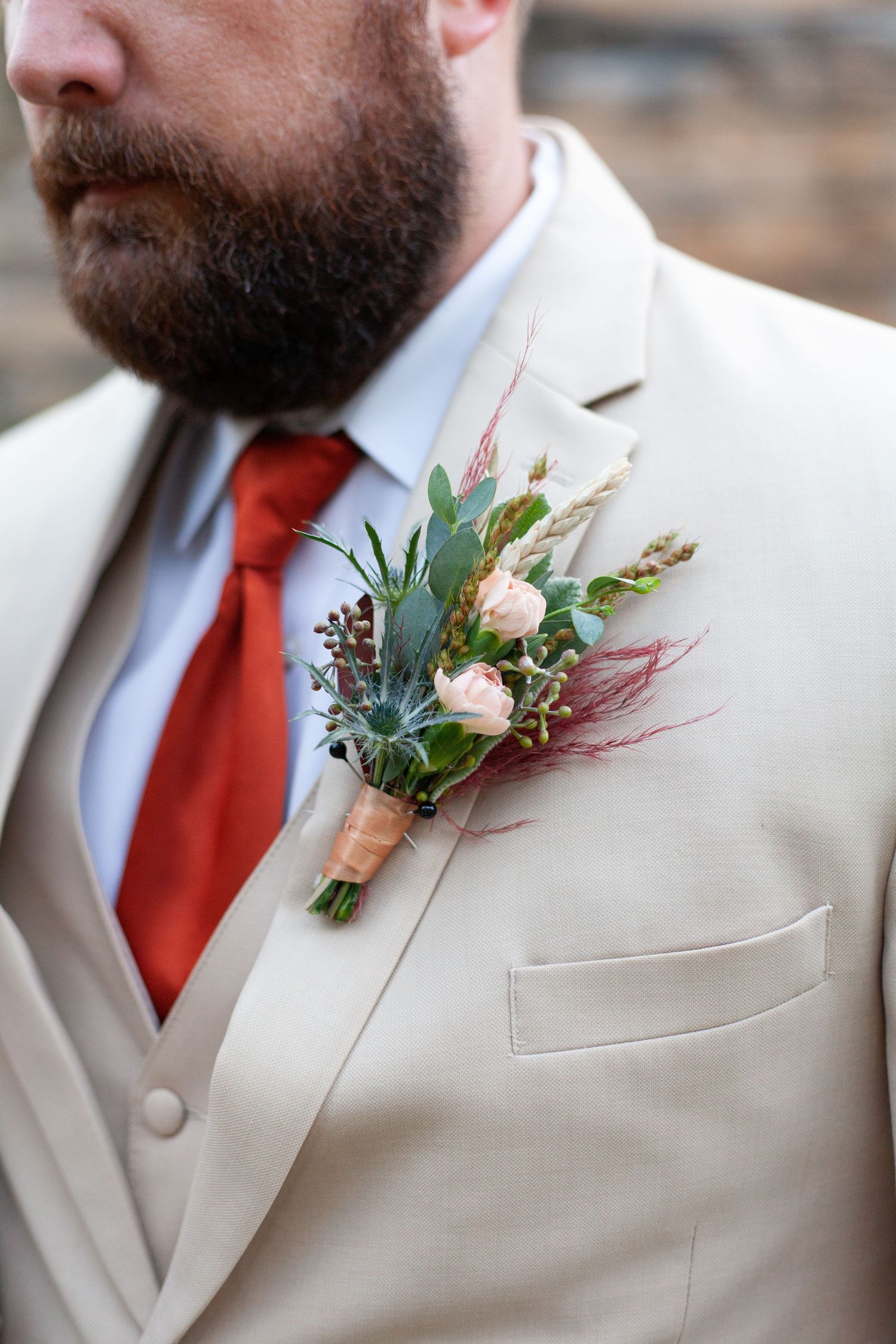 A delicate floral boutonniere with pink roses and eucalyptus adorns a tan suit with red tie.