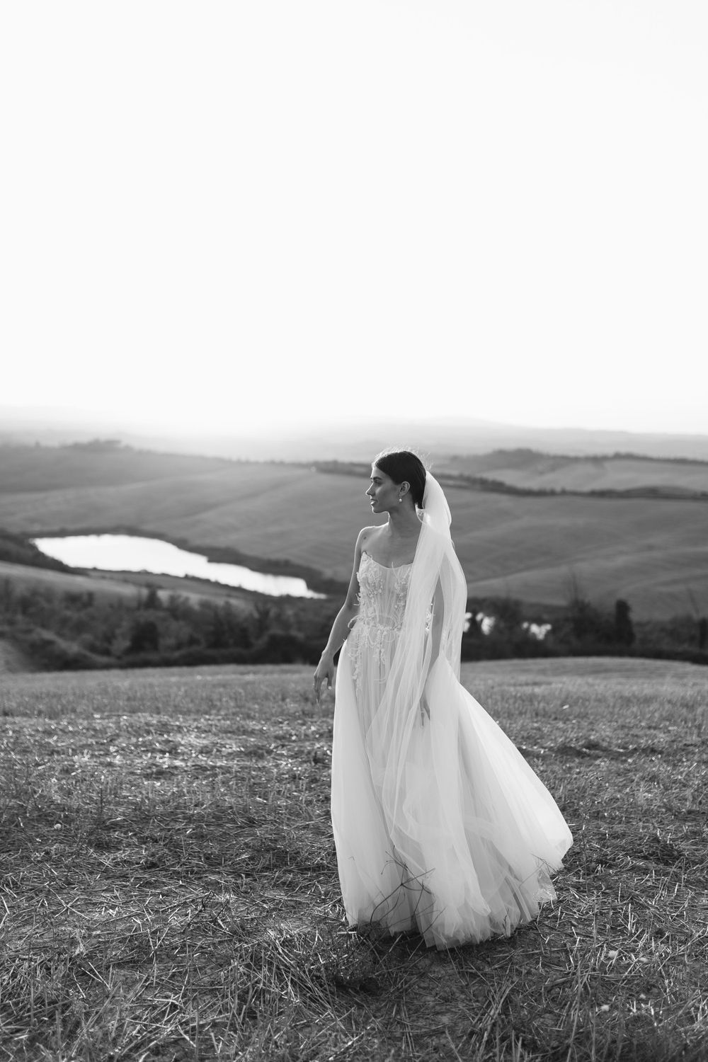 Black and white photo of a bride in a flowing gown standing on a hilltop with lake view.
