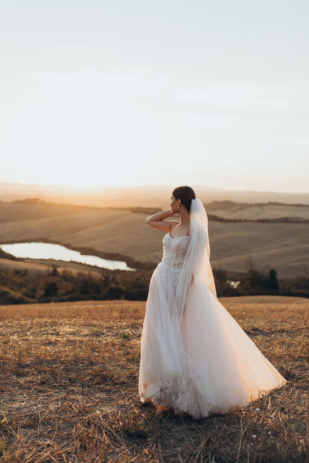 Elegant bride in lace gown with flowing veil silhouetted against sunset mountains.