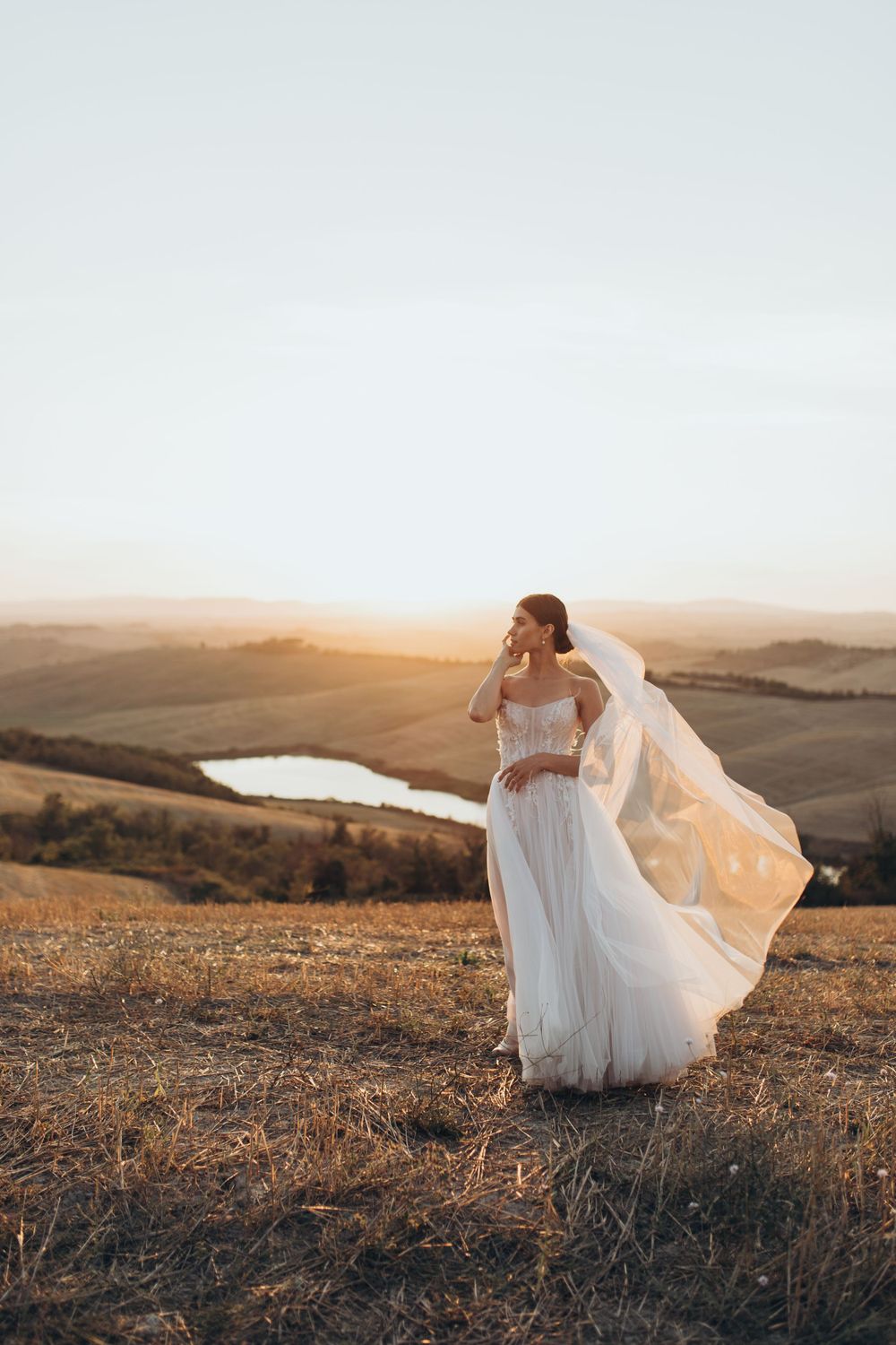 Wedding dress and veil catch golden light on hilltop overlooking countryside with distant lake at sunset.