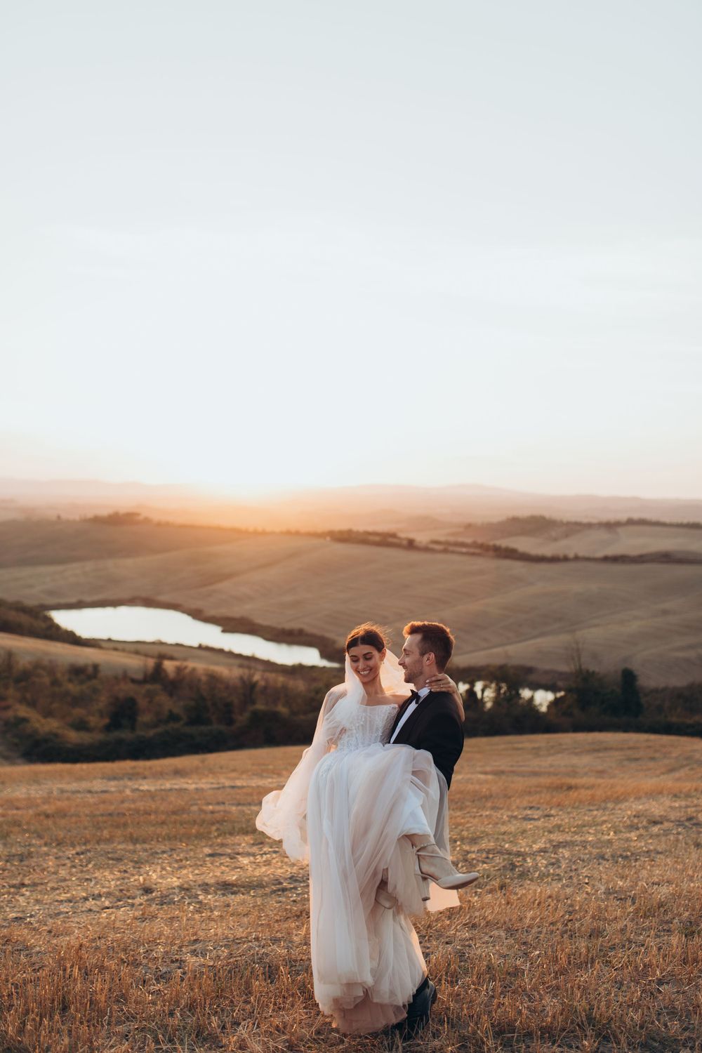 A newlywed couple share a romantic moment on a hilltop during golden hour with a lake in the background.
