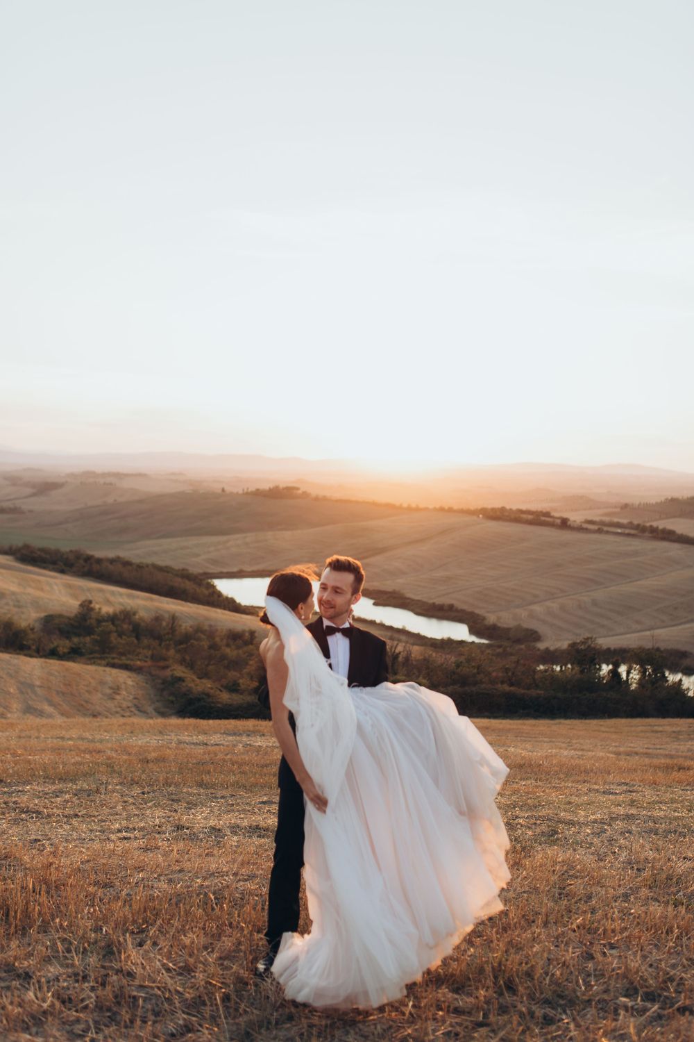 A couple in wedding attire pose dramatically on a hilltop during sunset with golden light.