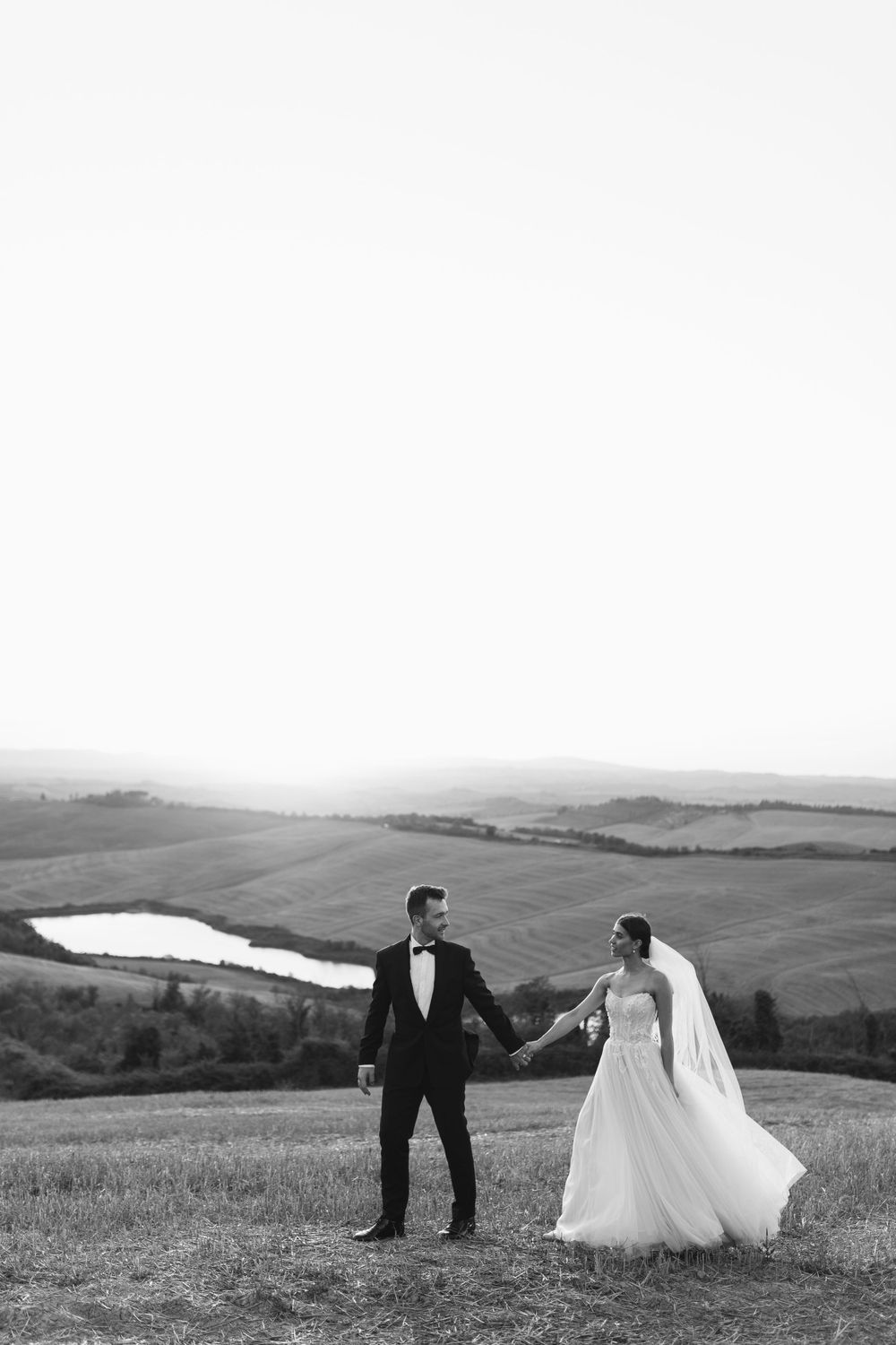 Black and white photograph of wedding couple holding hands on hilltop with countryside view.