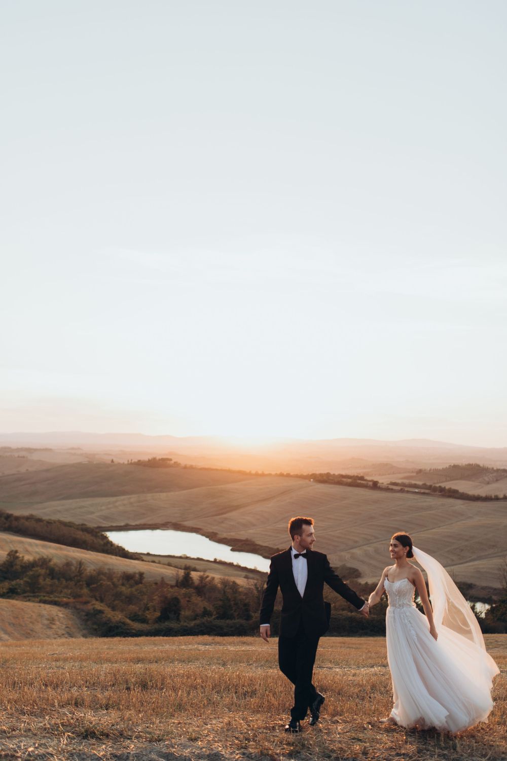 Wedding couple walks hand in hand on golden hillside during sunset with lake in distance.