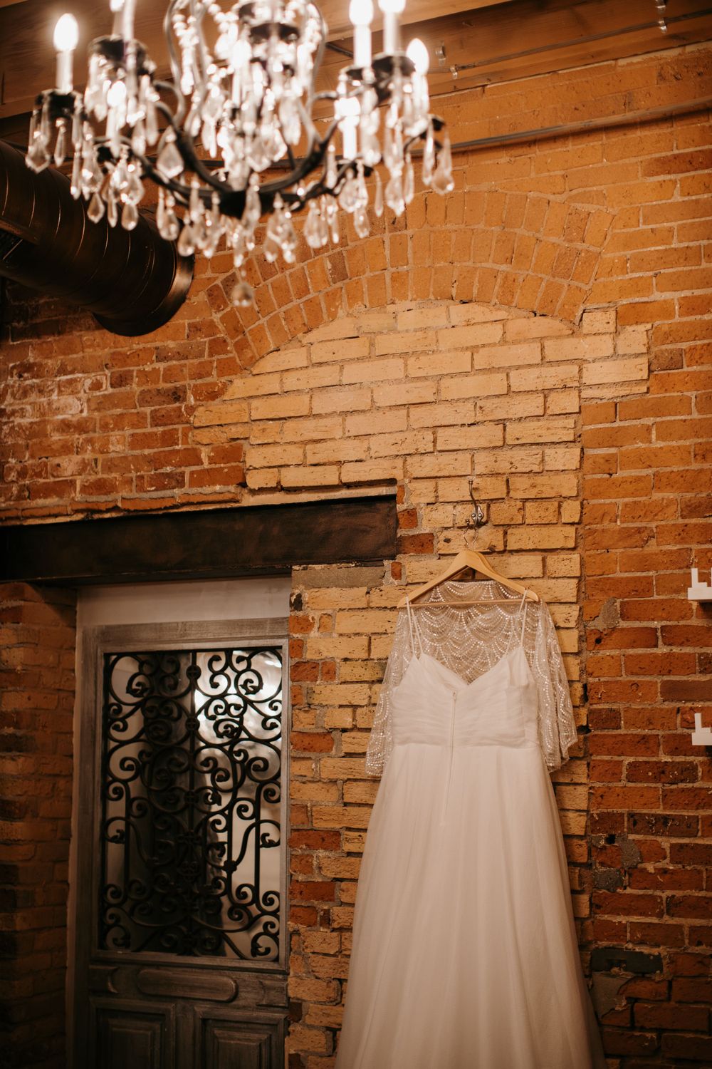 White lace wedding dress hanging near a crystal chandelier against a rustic exposed brick wall.