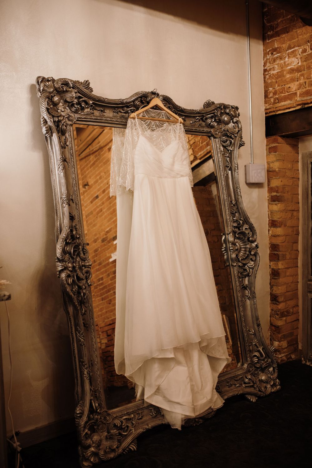 Elegant white wedding dress with lace cape overlay hanging on an ornate silver mirror frame.