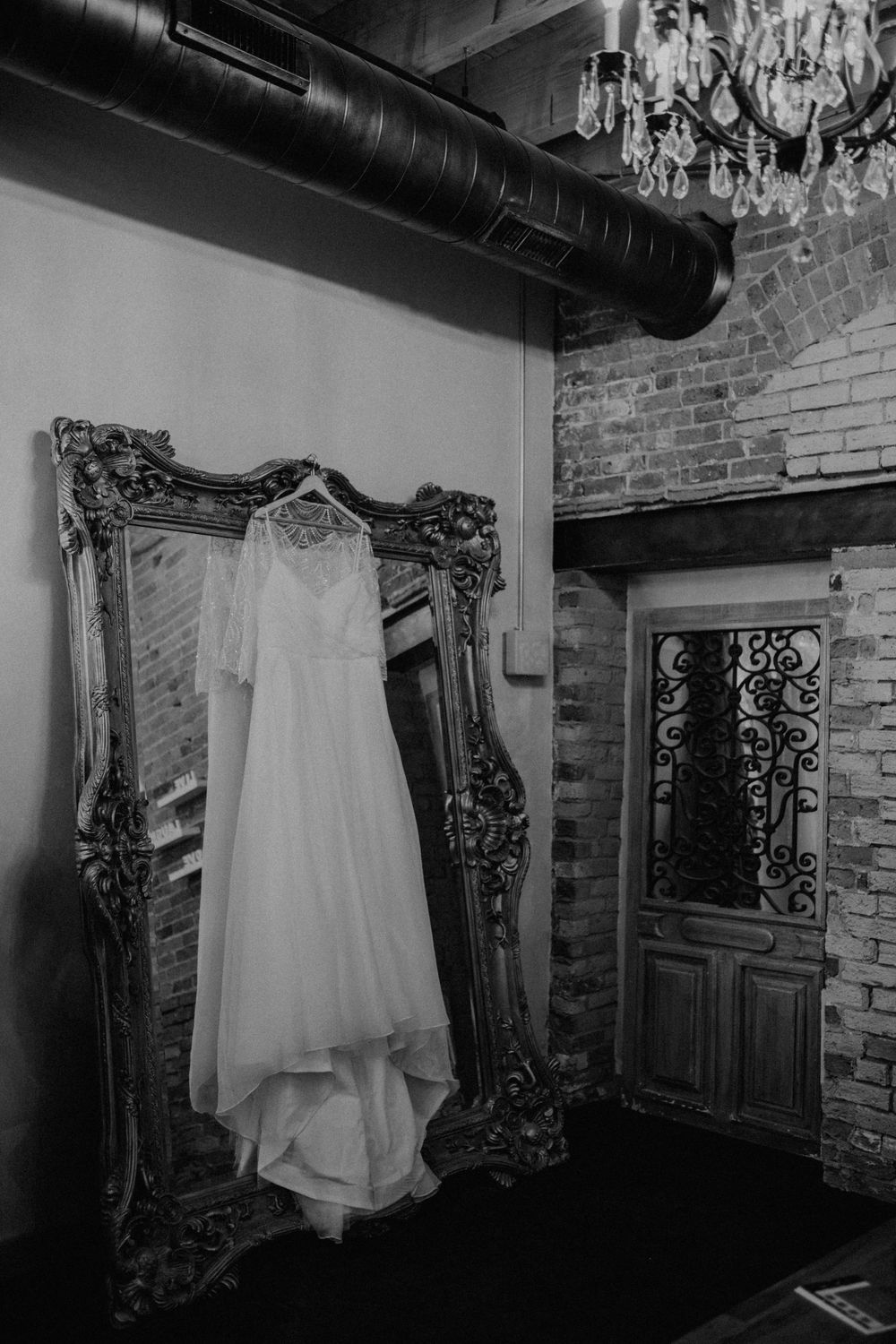 Black and white photo of a wedding dress hanging on an ornate mirror in a rustic brick venue.