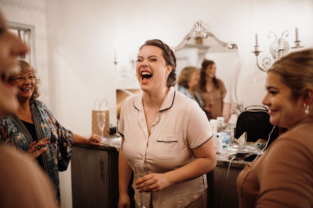 Joyful bride laughing with bridesmaids during wedding morning preparations in a bright room.