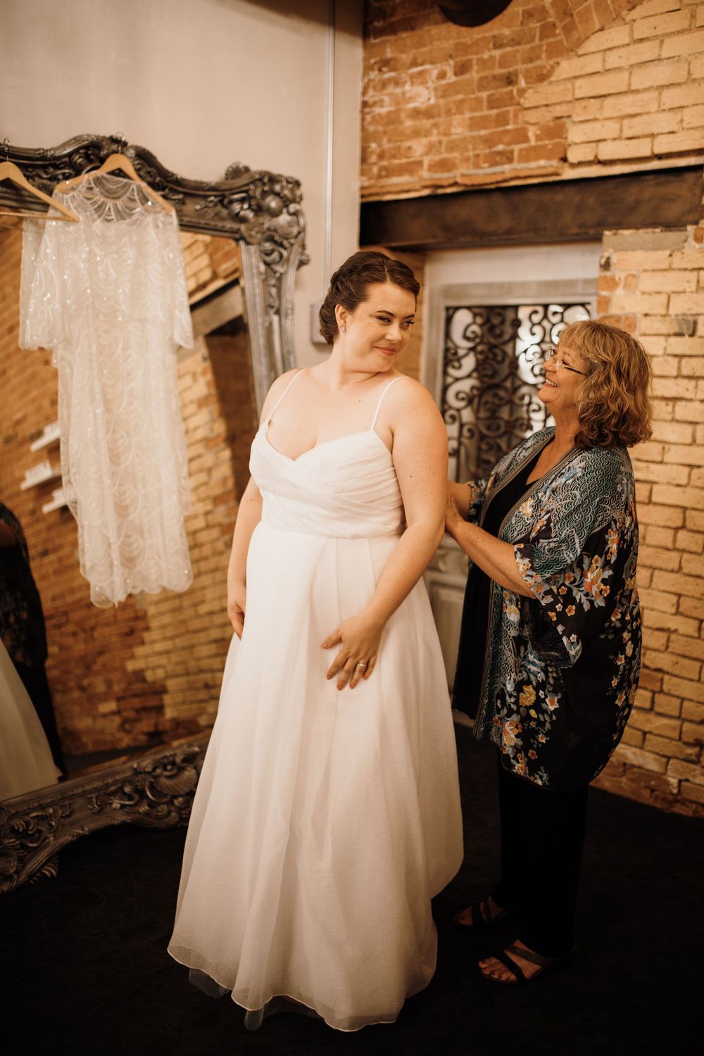 Mother helping bride adjust her white wedding dress in a rustic brick-walled bridal suite.