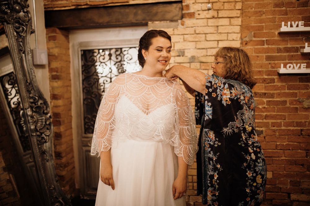 Mother placing lace capelet on bride's shoulders in a charming rustic brick wedding venue.