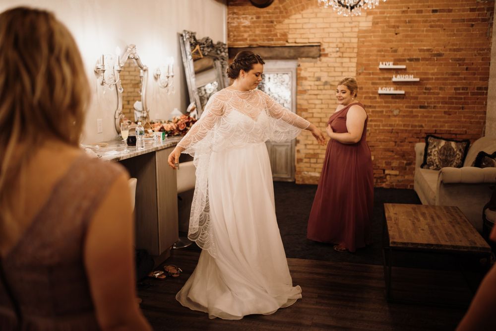 Bridesmaid in mauve dress helps bride adjust her elegant lace wedding gown indoors.