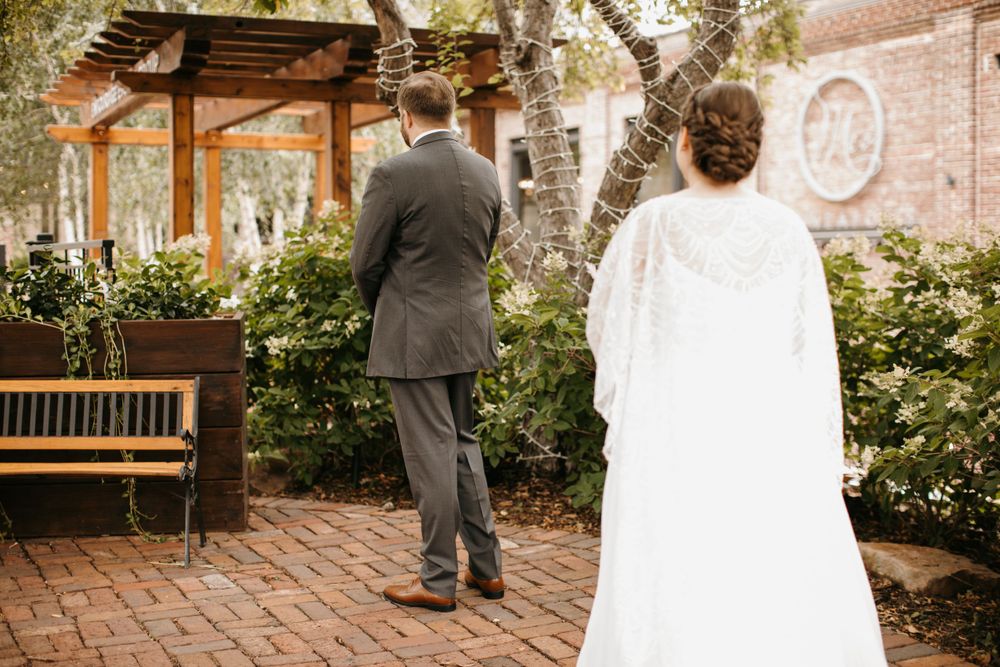 Bride in lace cape gown approaches groom from behind for their first look on brick path.