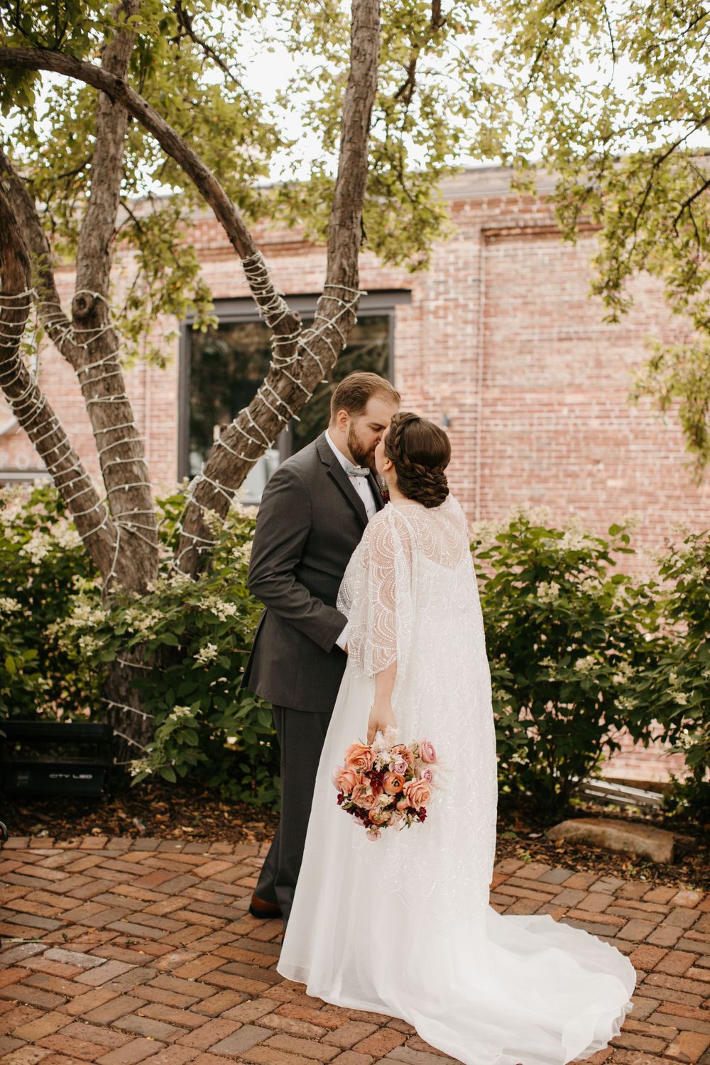 Bride and groom kiss tenderly in front of a brick building surrounded by blooming greenery.