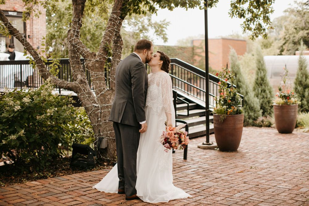 Bride and groom share a romantic kiss outdoors on a brick patio surrounded by trees.
