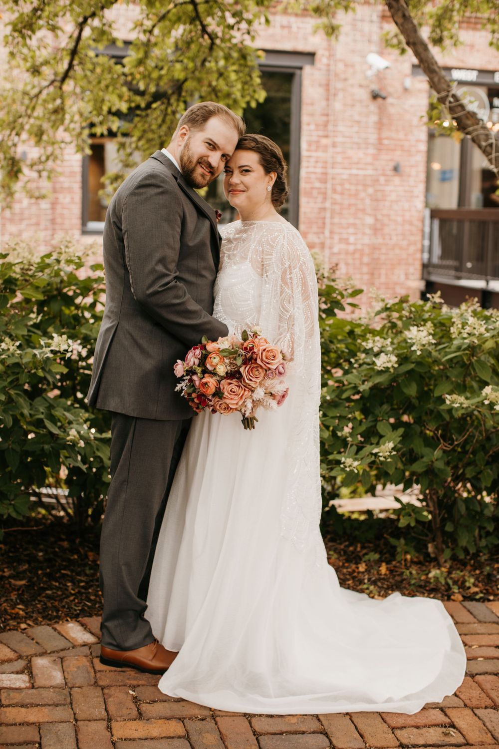 Bride and groom pose together smiling holding a pink and coral bouquet near brick building.