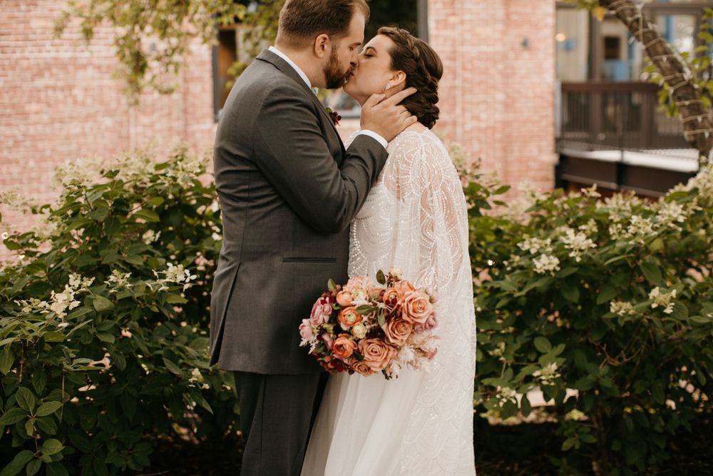Bride and groom share a passionate kiss surrounded by lush white flowering greenery.