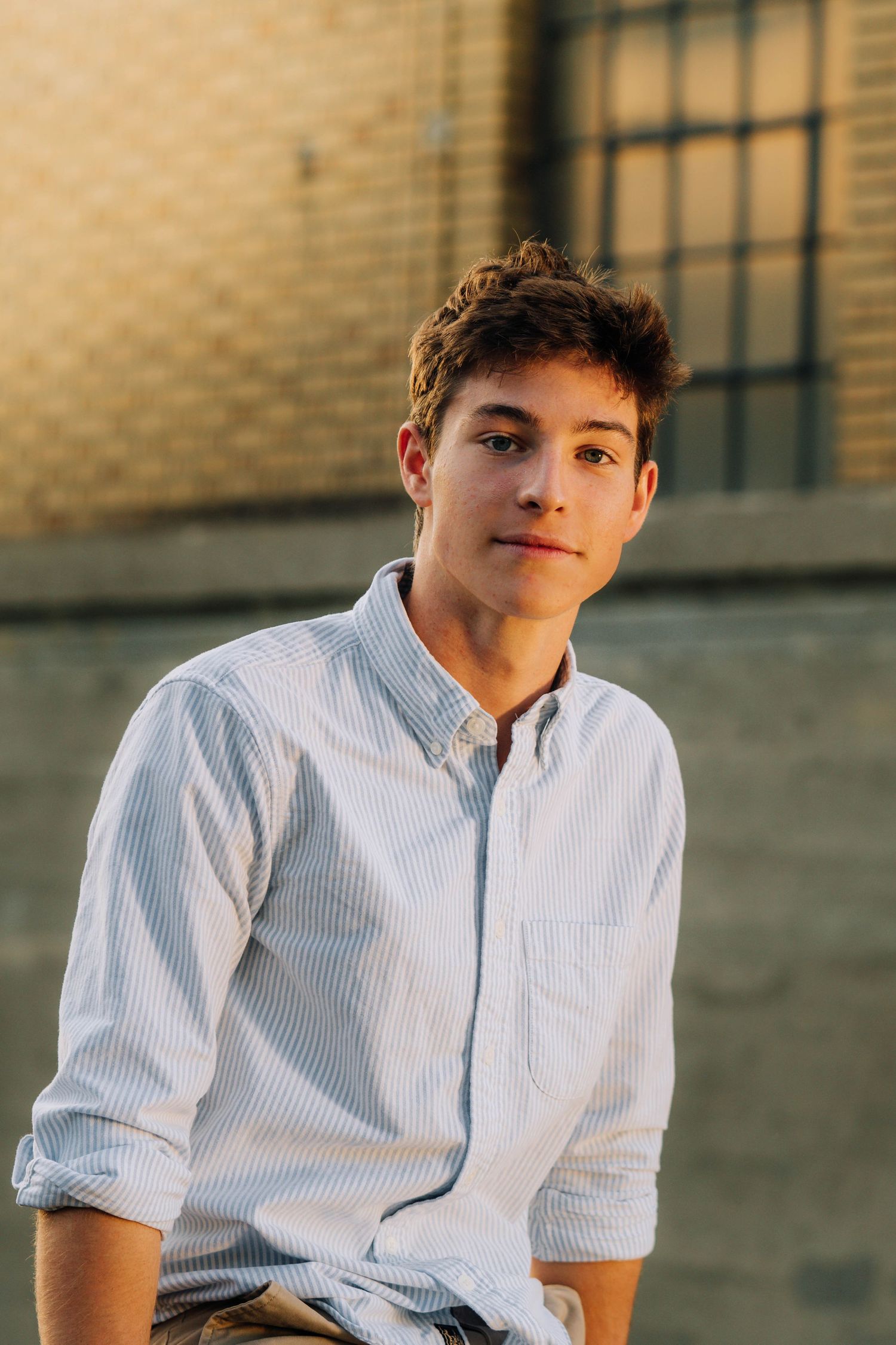 Portrait of someone in striped button-down shirt sitting against brick wall at sunset.