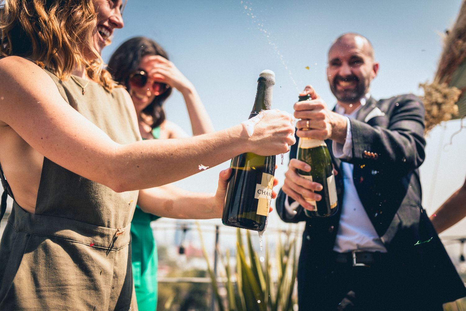 Wedding guests toasting with champagne bottles on a sunny outdoor terrace.