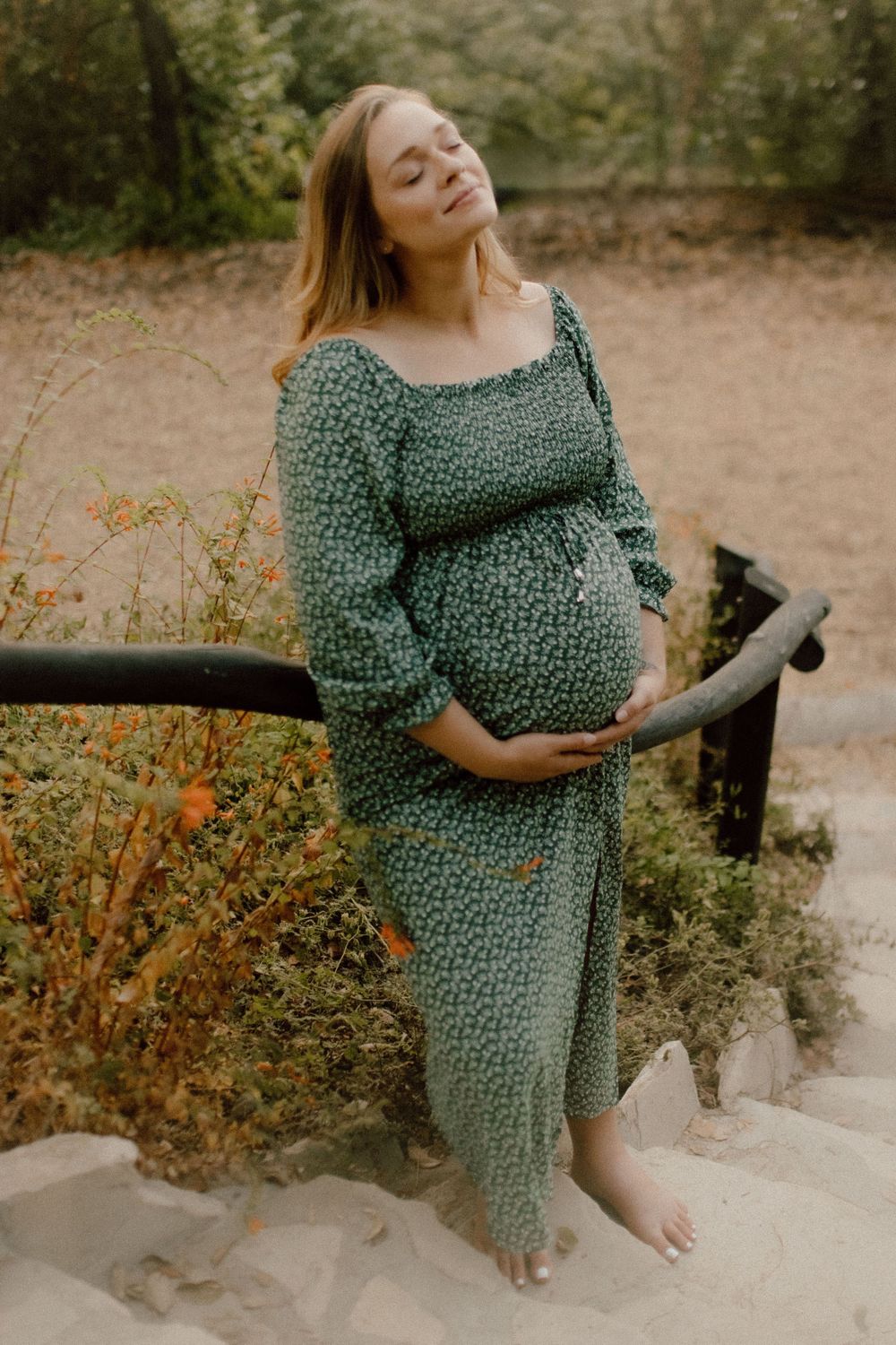 Pregnant woman in sage green floral dress poses barefoot on sandy ground during autumn outdoor maternity photoshoot.