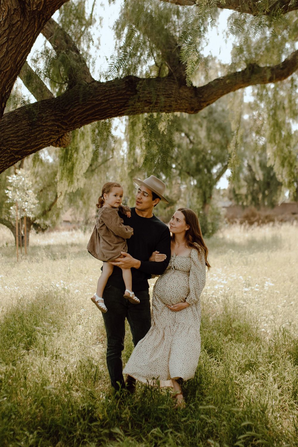 A couple stands beneath a willow tree in a dreamy meadow filled with wildflowers on a sunny afternoon.