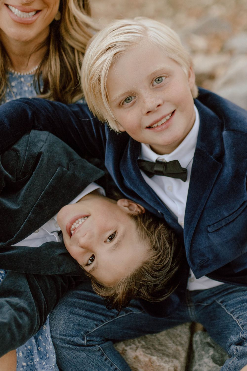 A playful family portrait with two children wearing formal navy blazers and bow ties laying on rocks outdoors.