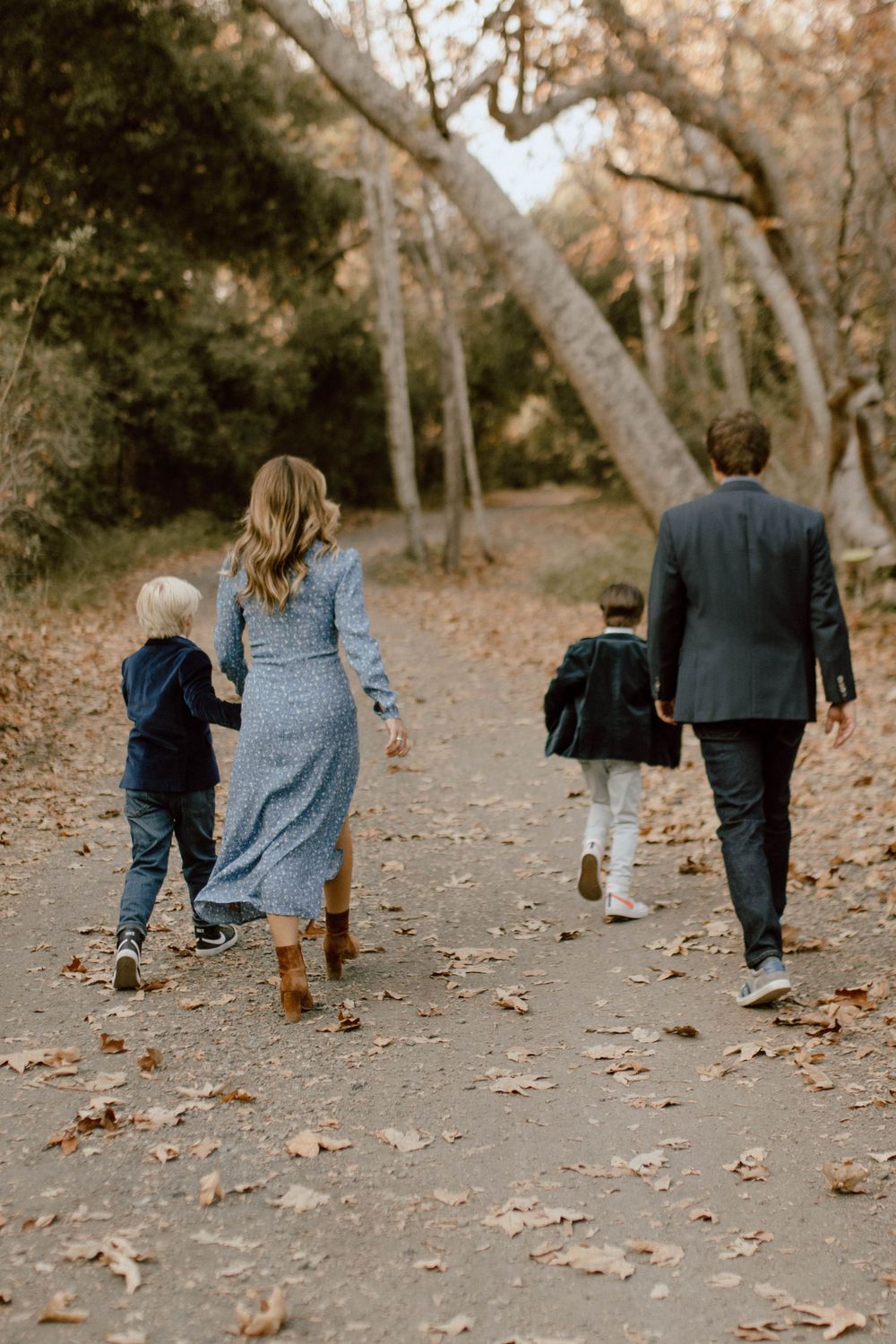 Two adults and two children walking together on a leaf-strewn forest path during autumn season.