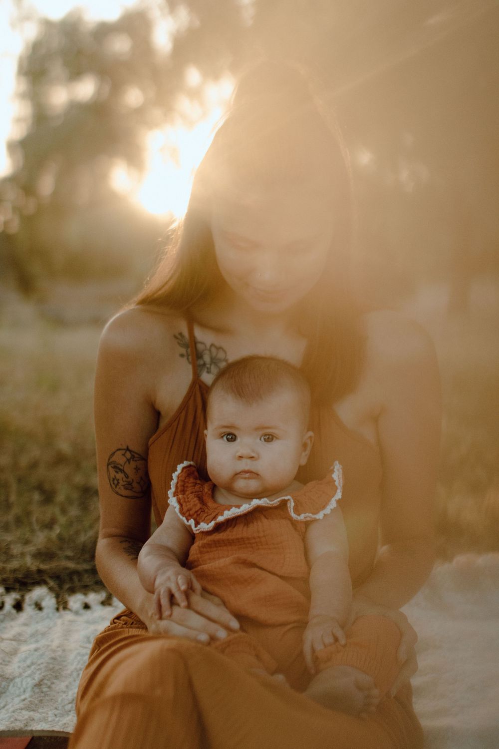 A parent in brown dress holds an infant wearing matching clothing during golden hour sunset outdoors.