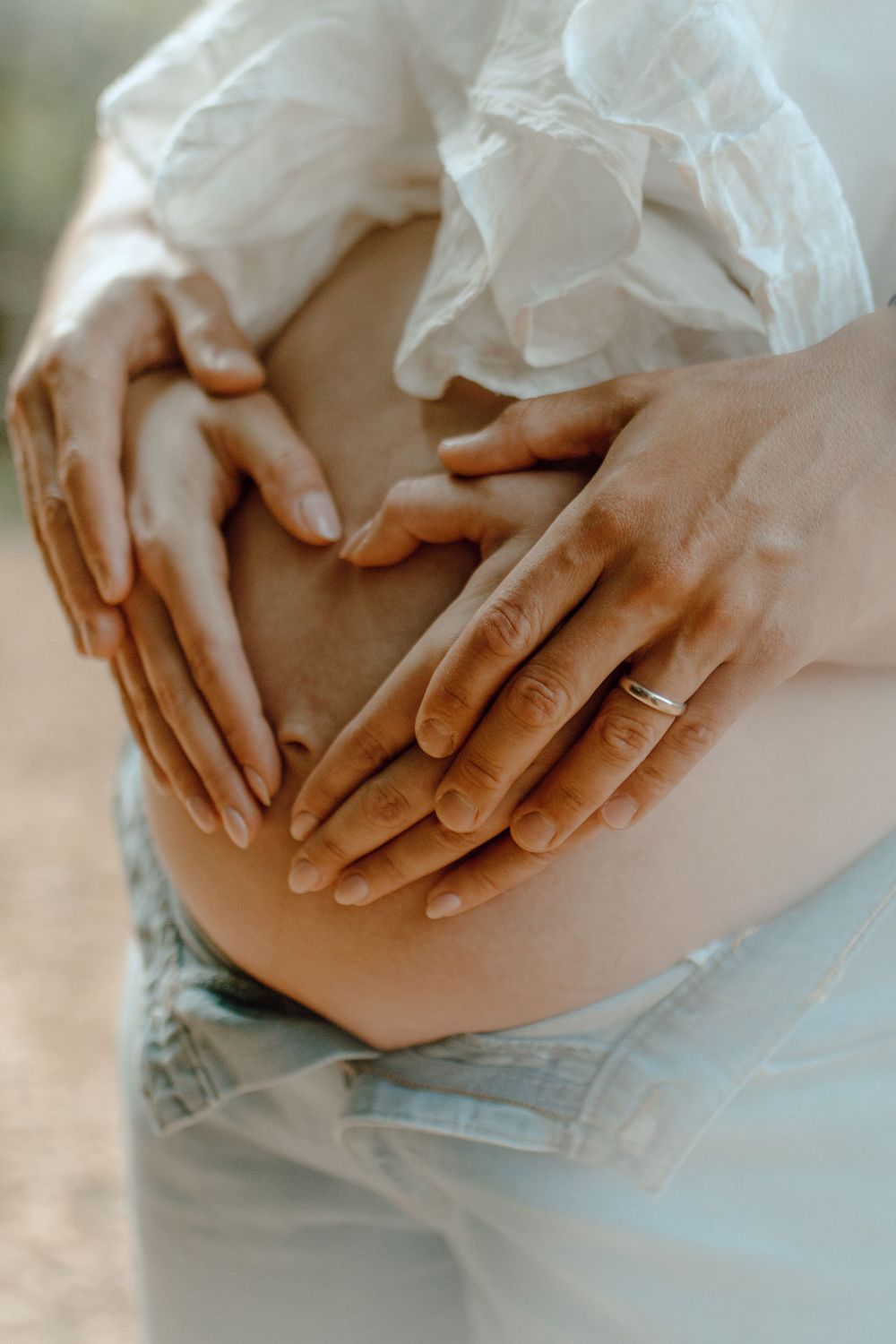 Close-up of hands forming a heart shape on a pregnant belly wearing a white flowy top and jeans.