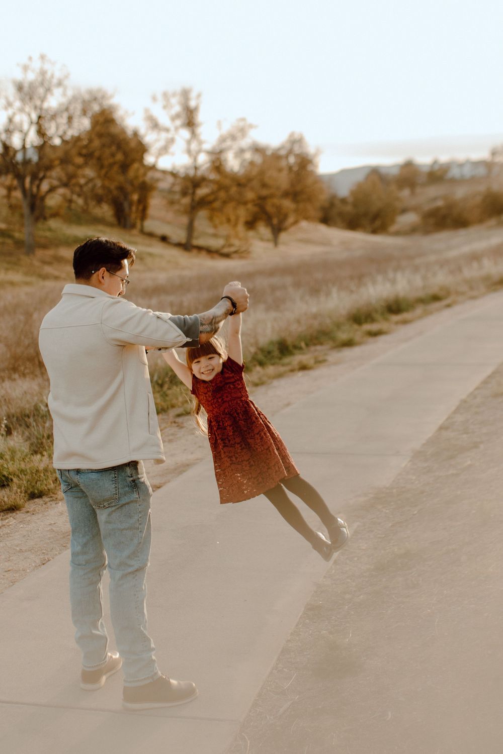 A parent spins a child in a red dress on a paved path surrounded by autumn trees and rolling hills at sunset.