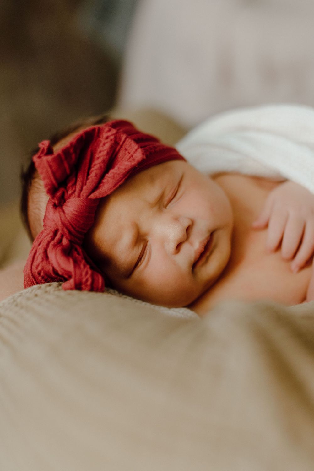 Peaceful sleeping newborn baby wearing a red bow headband rests on soft beige fabric.