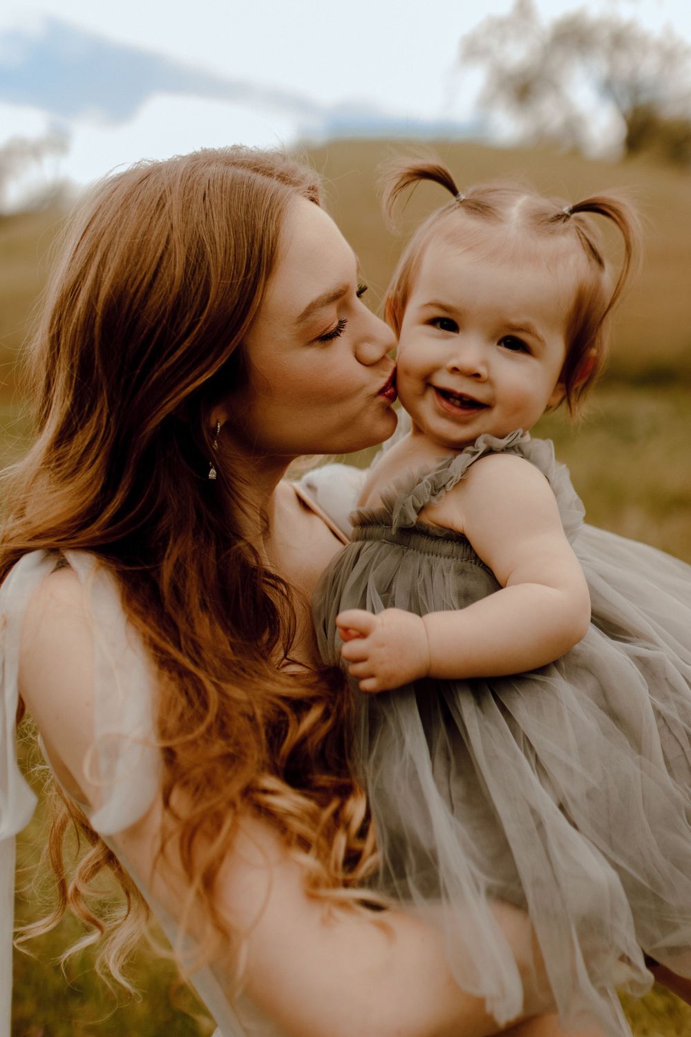 A tender moment between parent and child as they embrace outdoors in golden sunlight while the toddler wears a gray tulle dress.