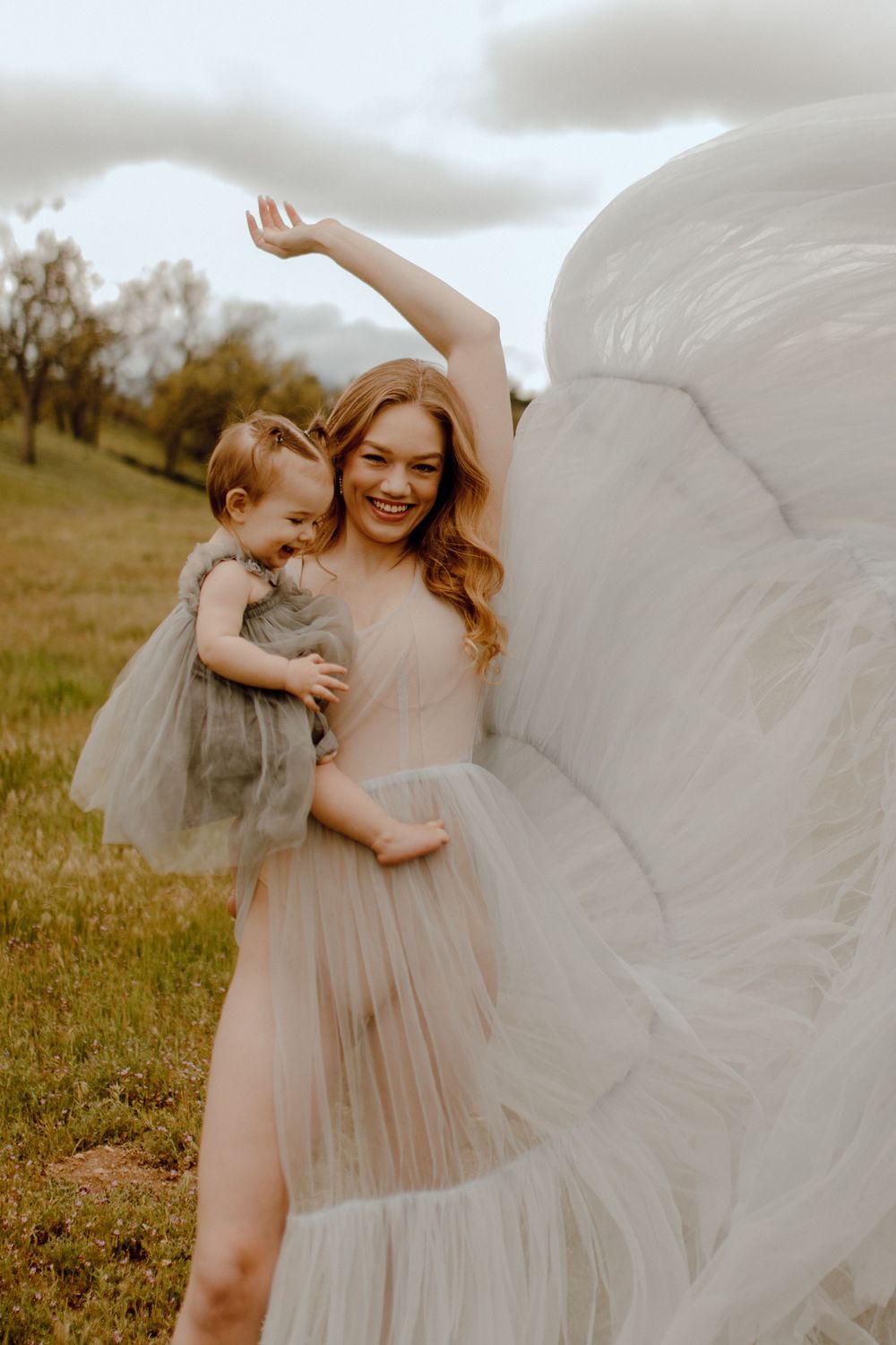 A joyful portrait in a field shows an intimate moment between a parent and child wearing flowy tulle dresses at sunset.