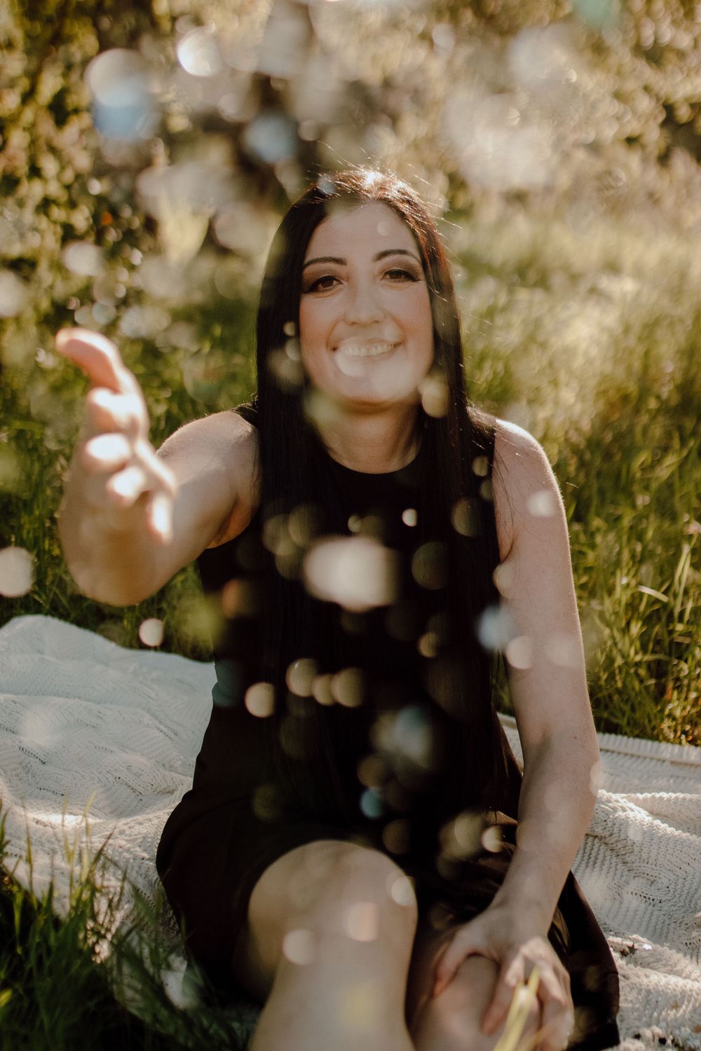 A person in a black dress sits on a blanket outdoors while bokeh effects create dreamy light spots in the sunlit scene.