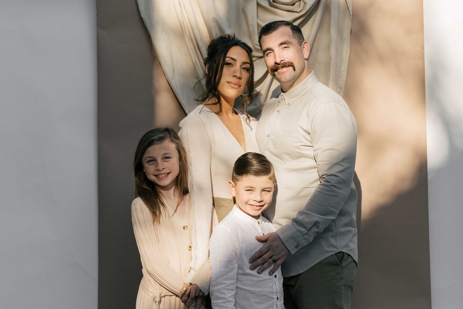 A happy family of four poses together in coordinating neutral outfits for a professional portrait session.
