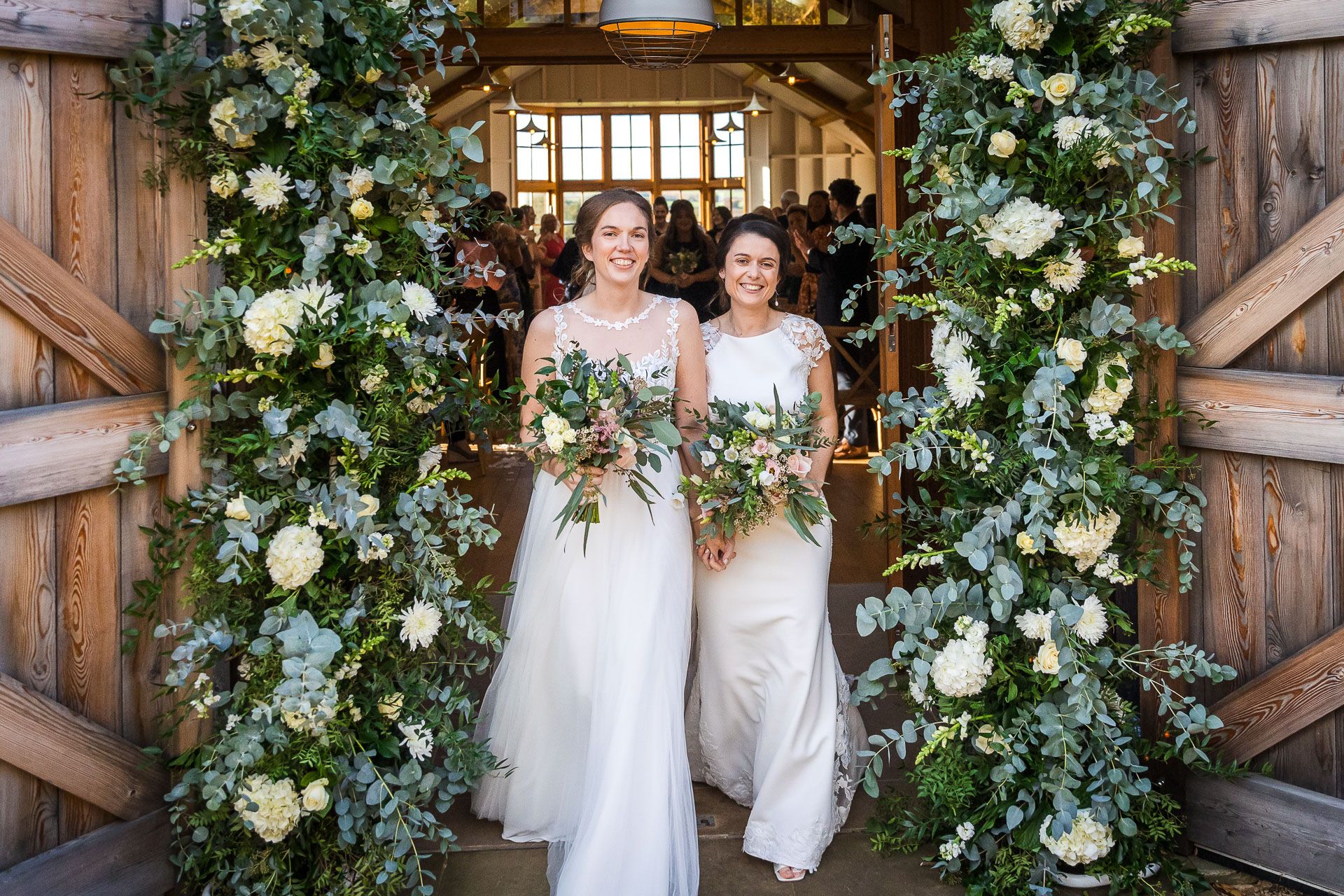 Two brides exit through rustic barn doors decorated with lush white floral arrangements and greenery.