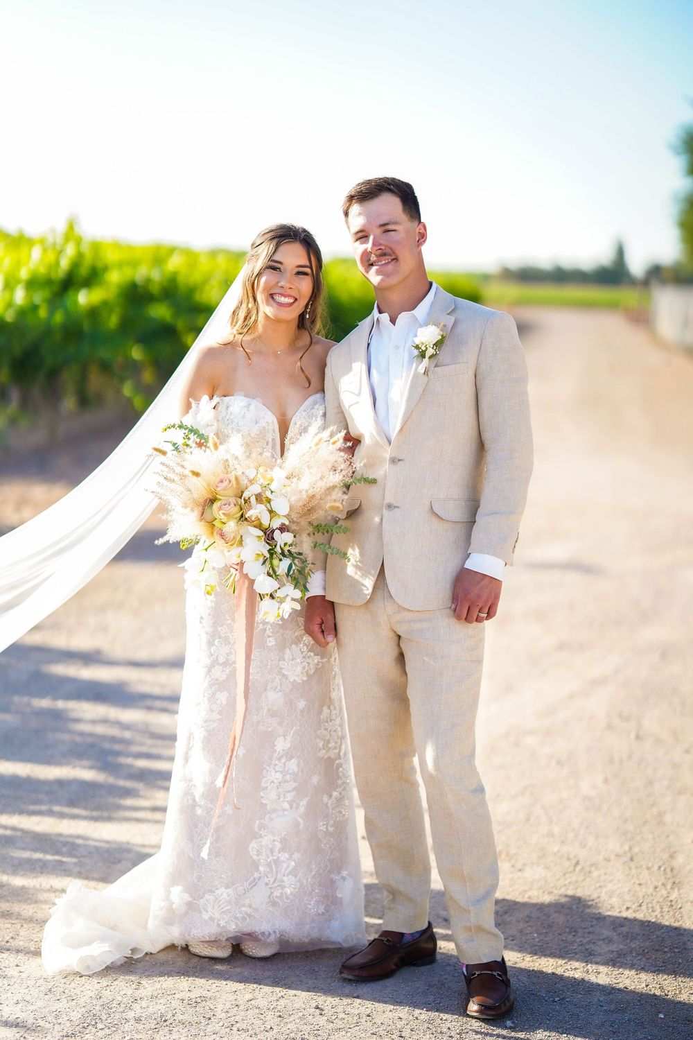 Bride in lace gown and groom in light suit posing together in vineyard setting.