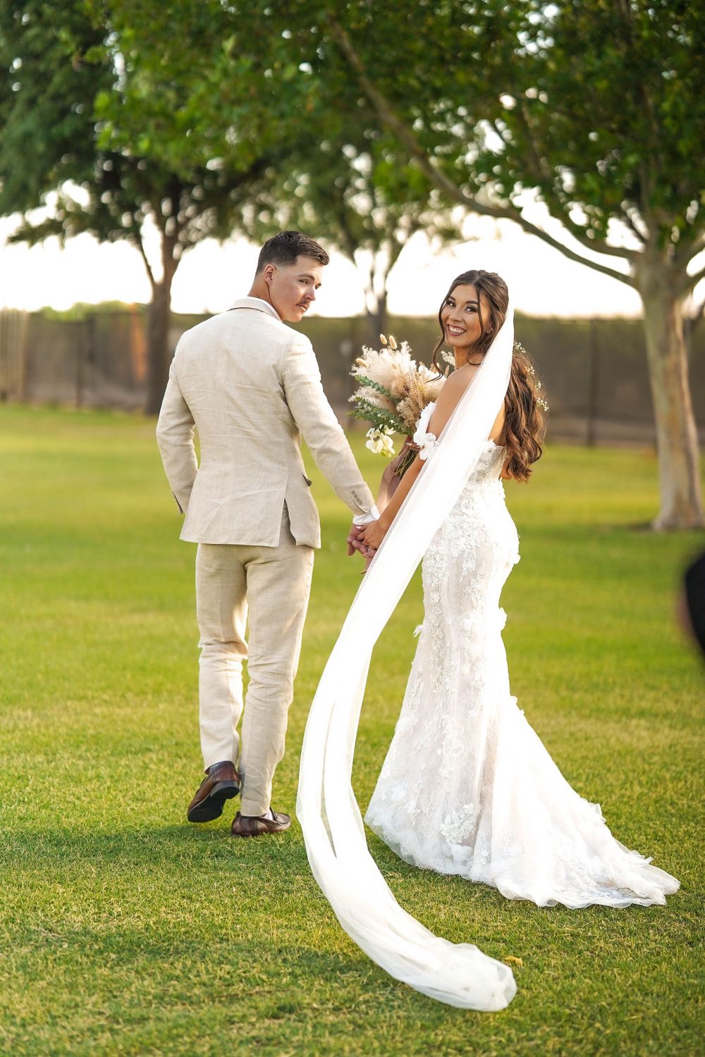 A wedding couple shares a playful moment on a lawn with a flowing white veil trailing behind.