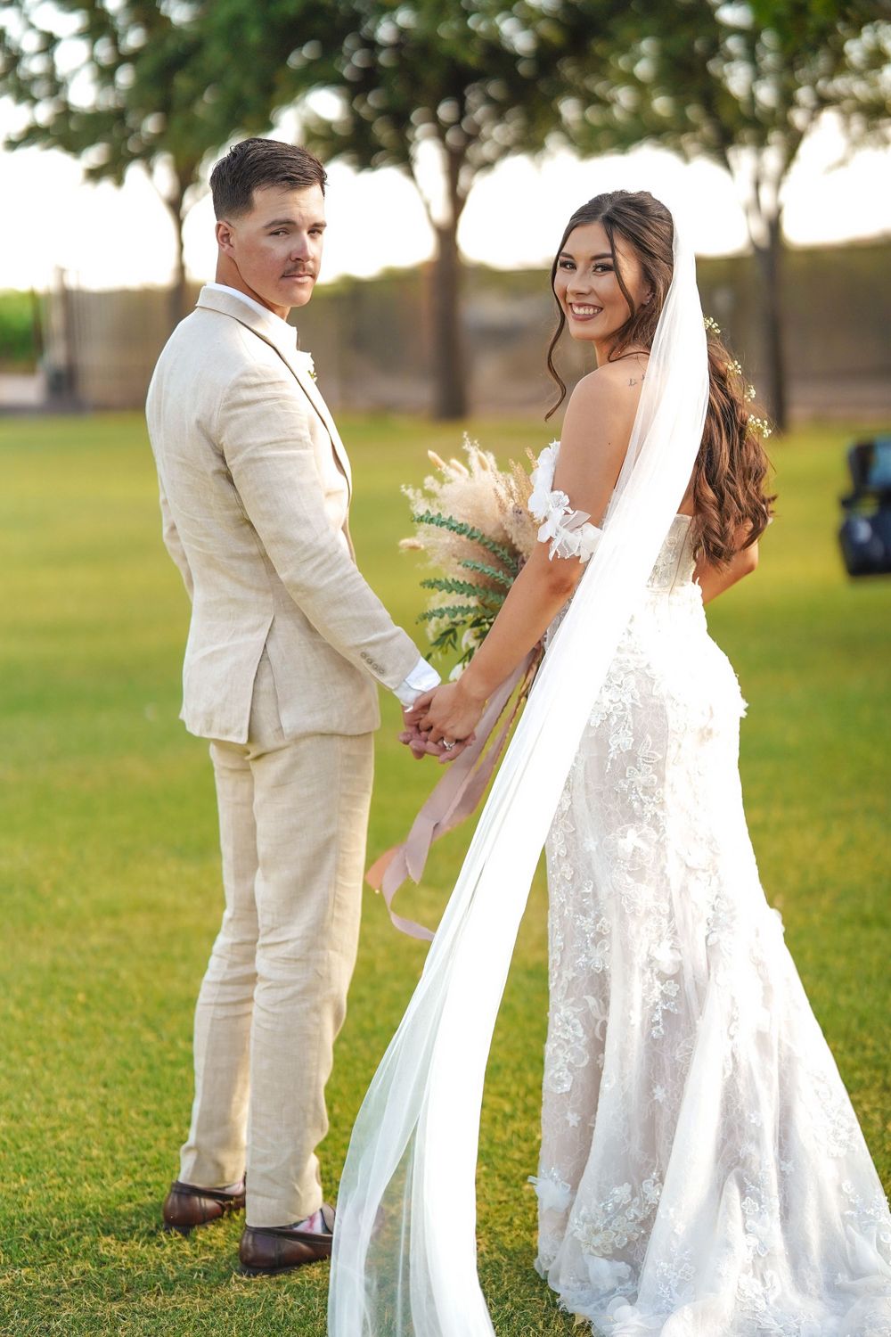 A couple in light-colored wedding attire poses on a grassy lawn with trees in the background.