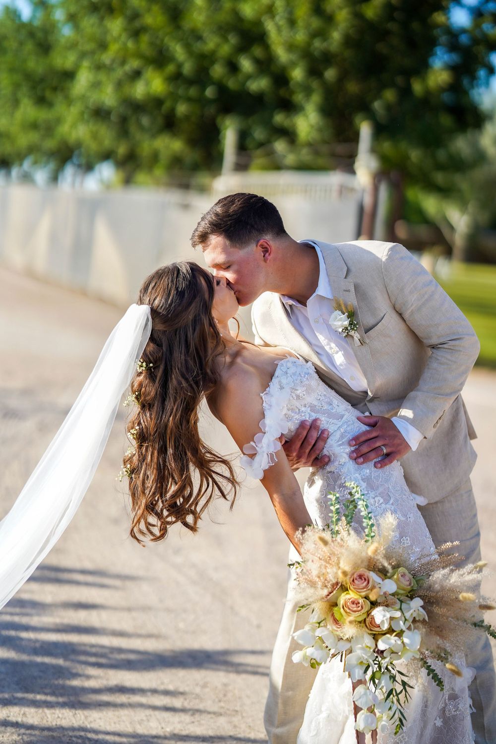 Romantic wedding couple sharing a tender dip kiss outdoors with a natural backdrop and beautiful floral bouquet.