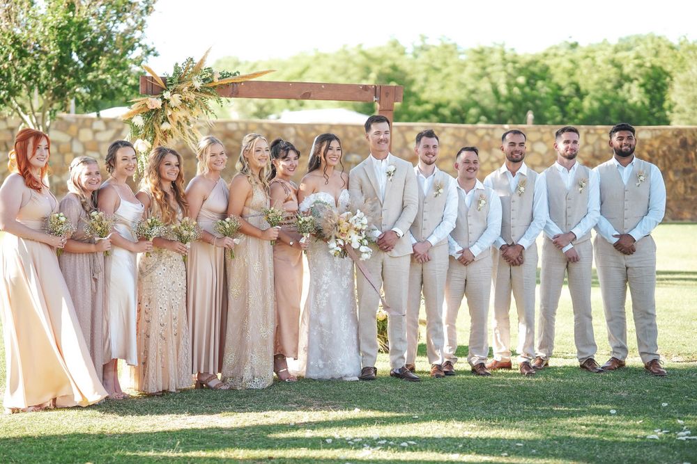 A wedding party in coordinated light-colored attire poses together on a sunny lawn.