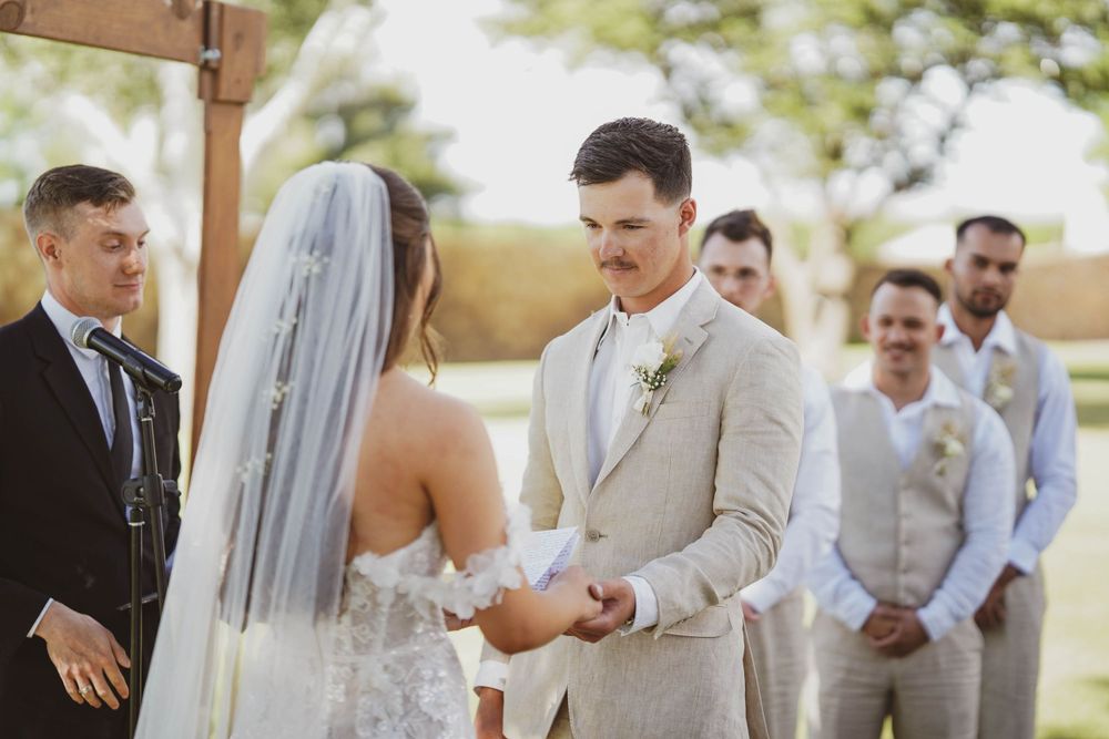 Couple exchanges rings during outdoor wedding ceremony under wooden arch with groomsmen watching.