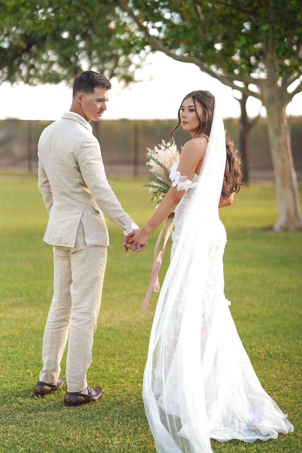 Playful bride and groom walking through a grassy garden setting in elegant white and cream attire.
