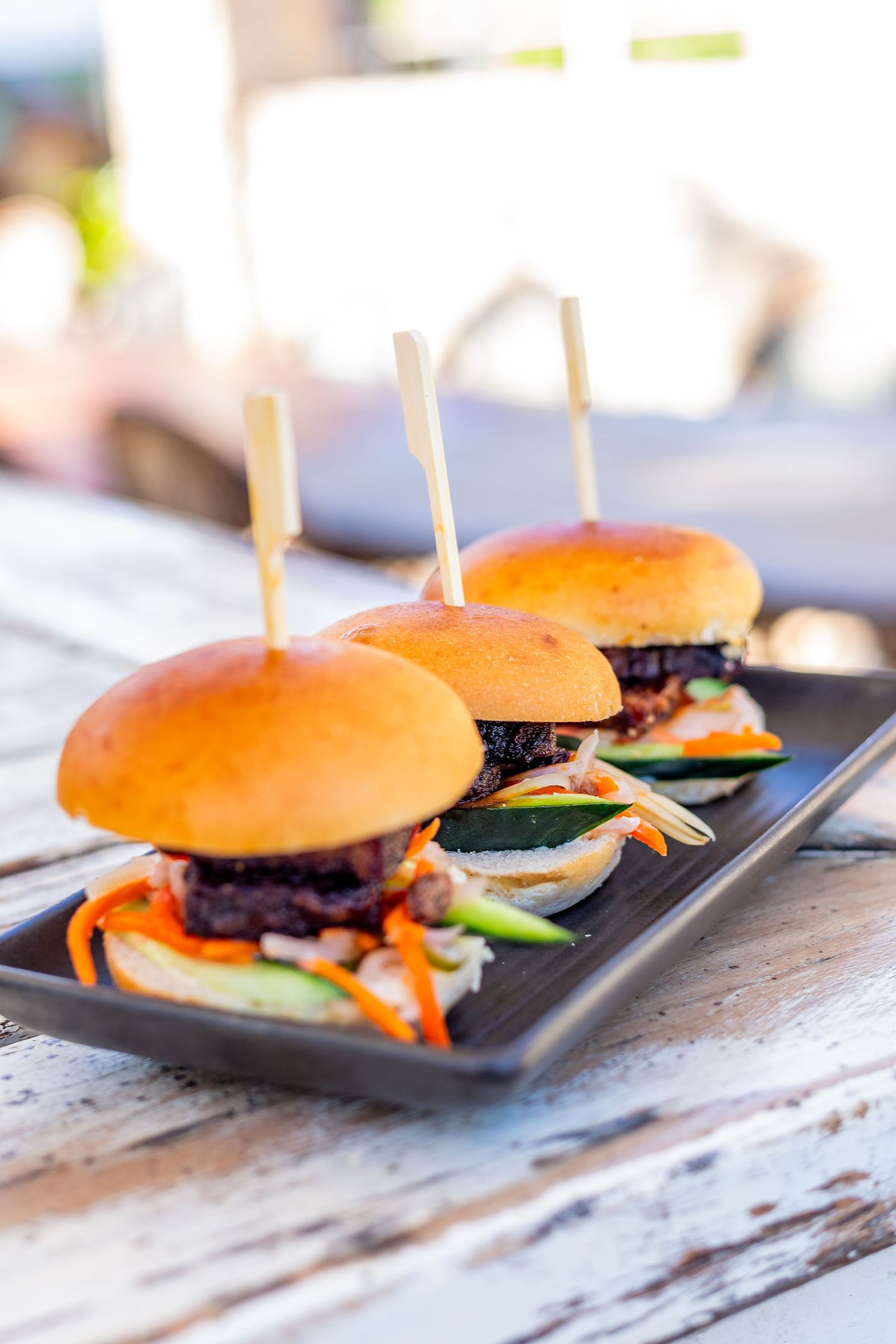 Three mini sliders with colorful garnishes served on black rectangular plate.