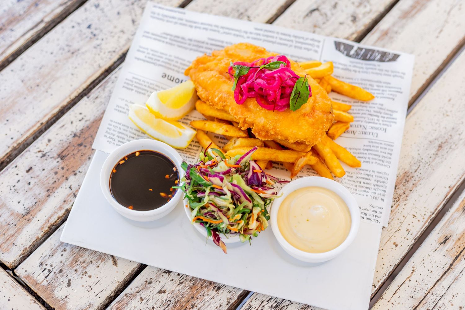 Crispy chicken fingers with french fries, coleslaw and two dipping sauces served on newspaper print paper.