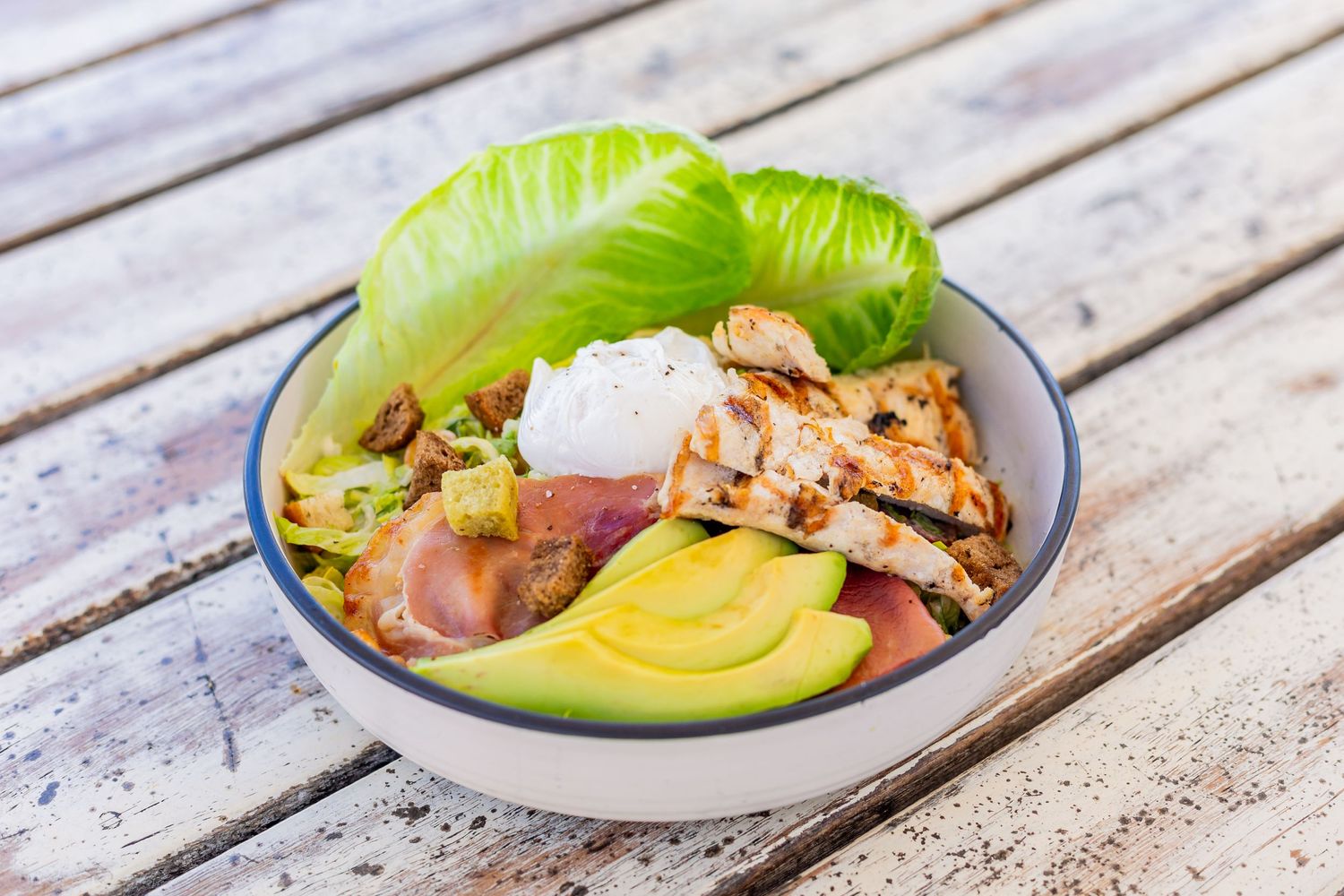 Fresh bowl with grilled chicken, avocado, lettuce and vegetables on a wooden surface.